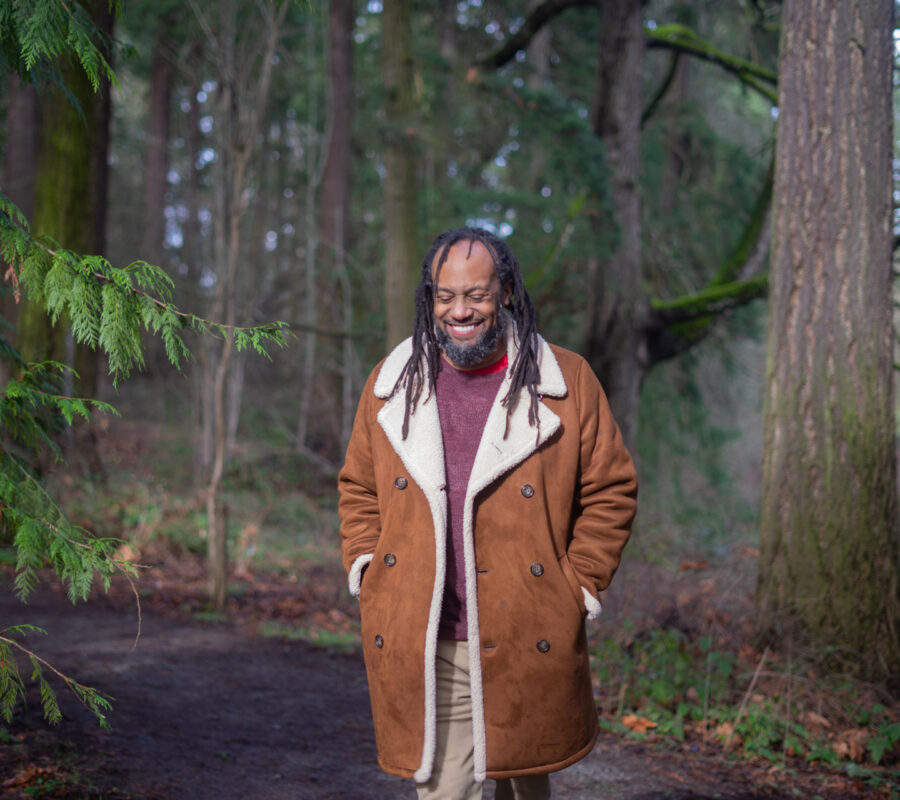 Tony wearing a brown and white coat smiling and standing in front of many green trees