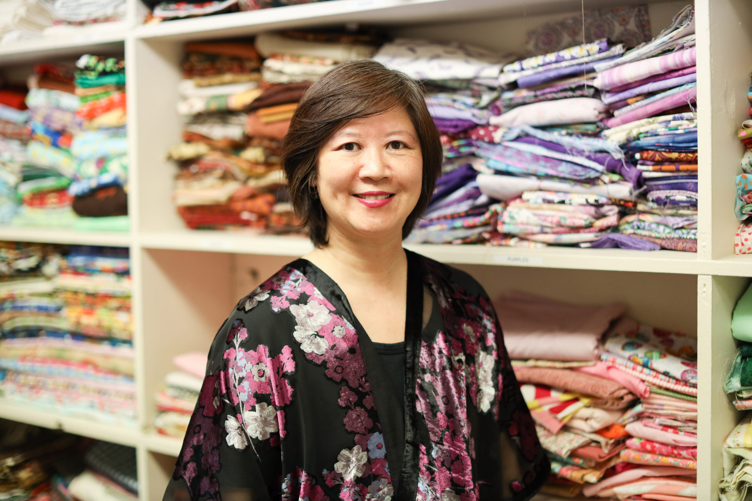 Ming-Ming wearing a black top with pink and white flowers standing in front of shelves of colored fabric.