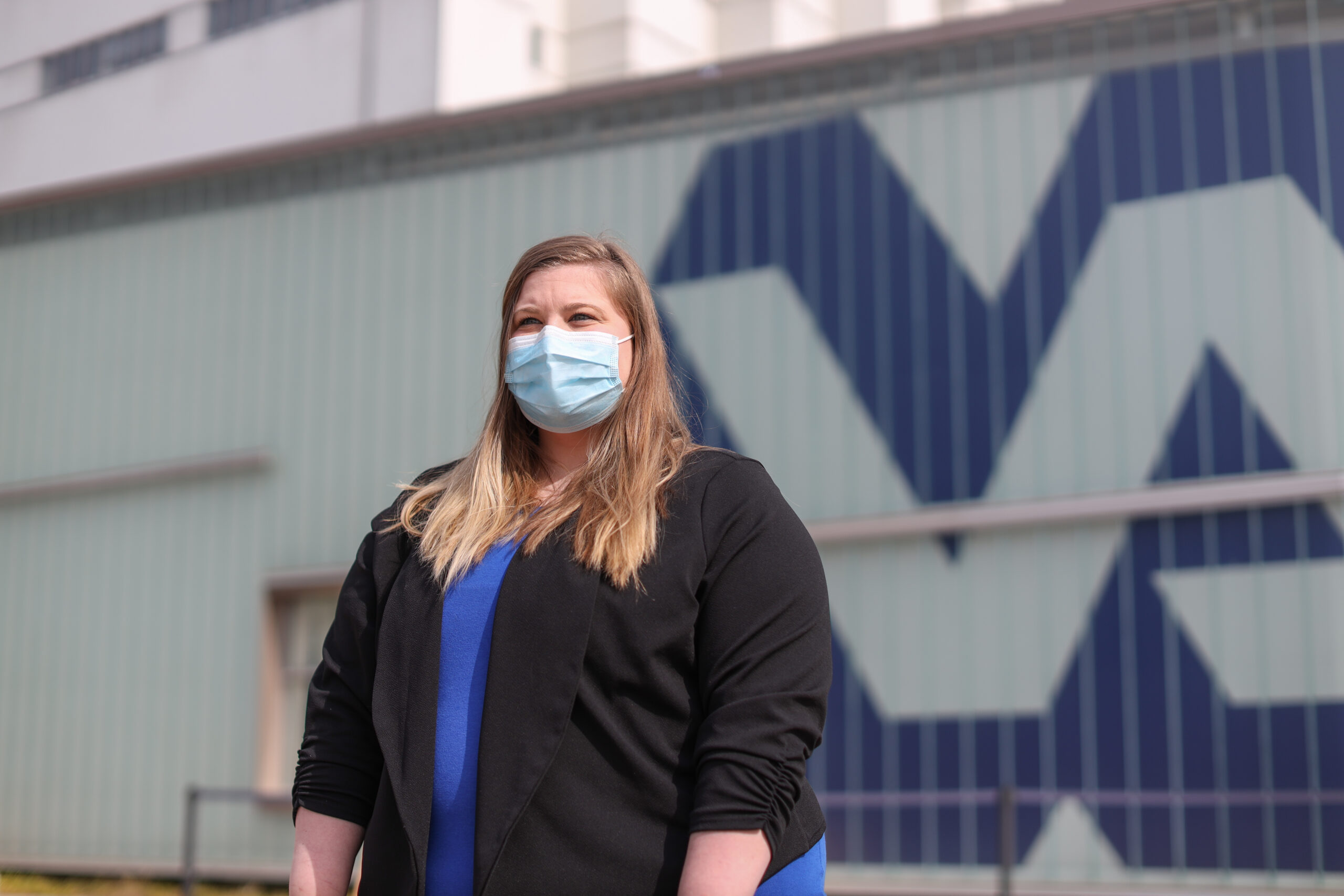 http://Katherine%20wearing%20a%20black%20sweater%20and%20blue%20shirt,%20and%20wearing%20a%20face%20mask%20standing%20outside%20with%20a%20building%20behind%20her