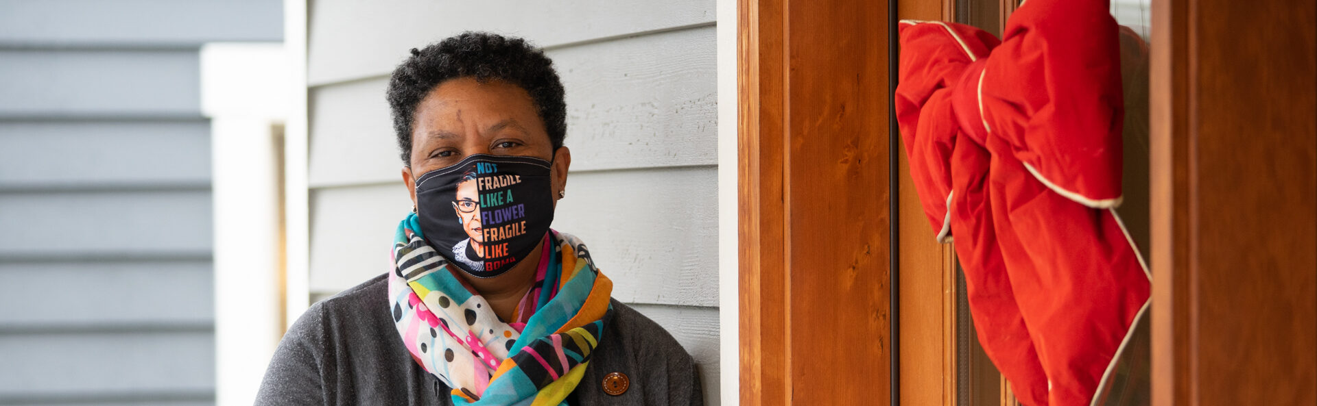 Lynda wearing a grey sweater, colorful scarf, and a face mask standing at a front door