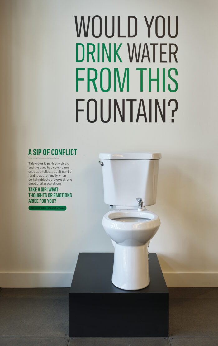 Image of a toilet drinking fountain with words on the wall behind it that says would you drink from this fountain?