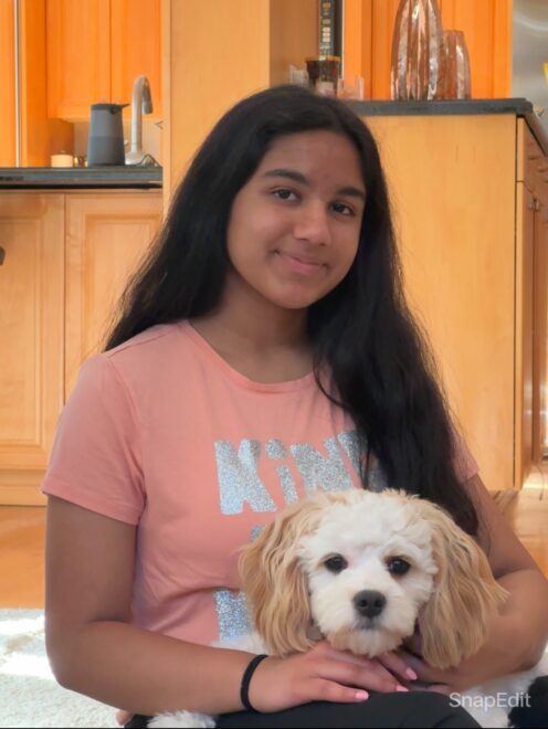 Vaanya wearing a pink shirt and holding a small white dog and smiling at the camera.