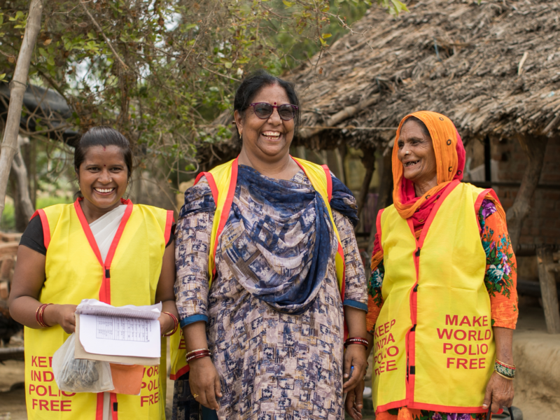 Three women sanding outdoors smiling wearing yellow polio vaccination vests.