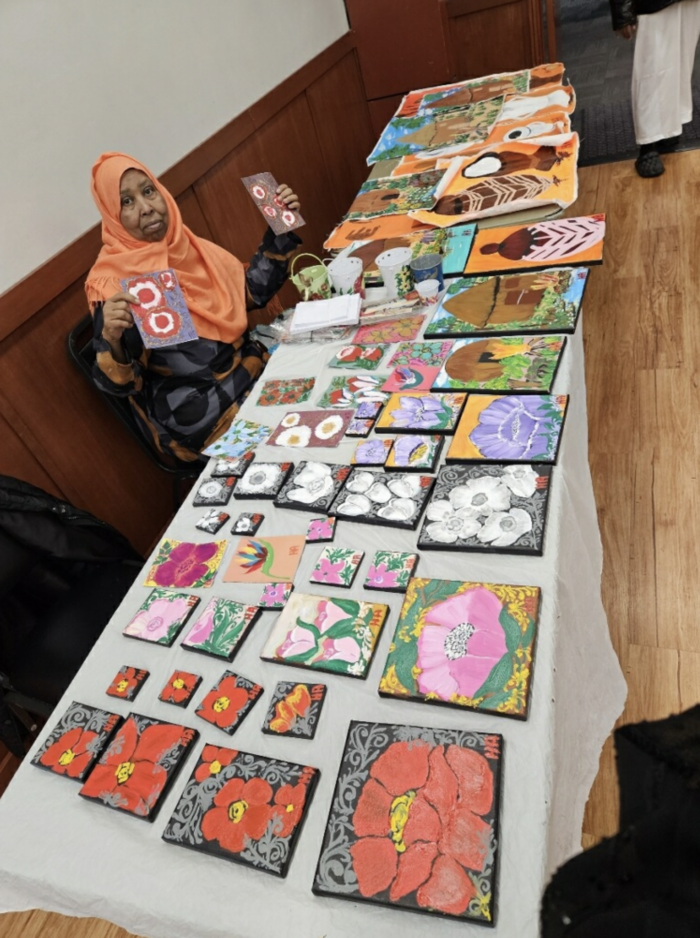 Hawo Arts artist sits at a table full of various art pieces for sale.